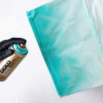 A black glove holds a can of Montana paint and sprays blue paint onto a cloth